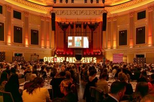 A large audience seated at round tables in an ornate hall watches a speaker on stage, with warm amber lighting, a large screen, and illuminated "QUEENSLAND GIVES" letters across the front of the stage.